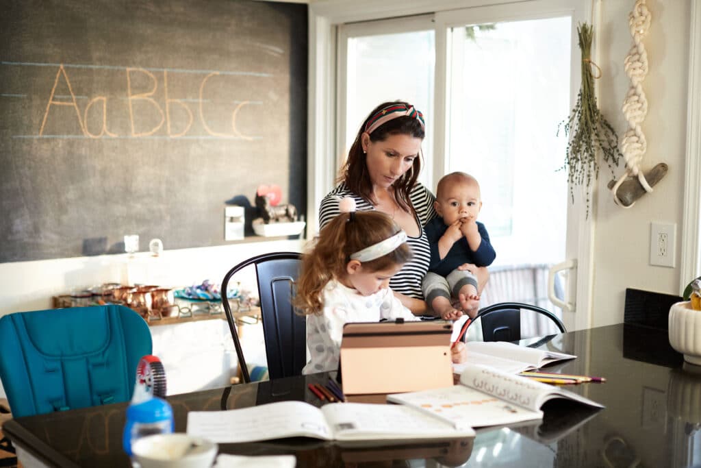 Mother holding an infant while homeschooling a young child