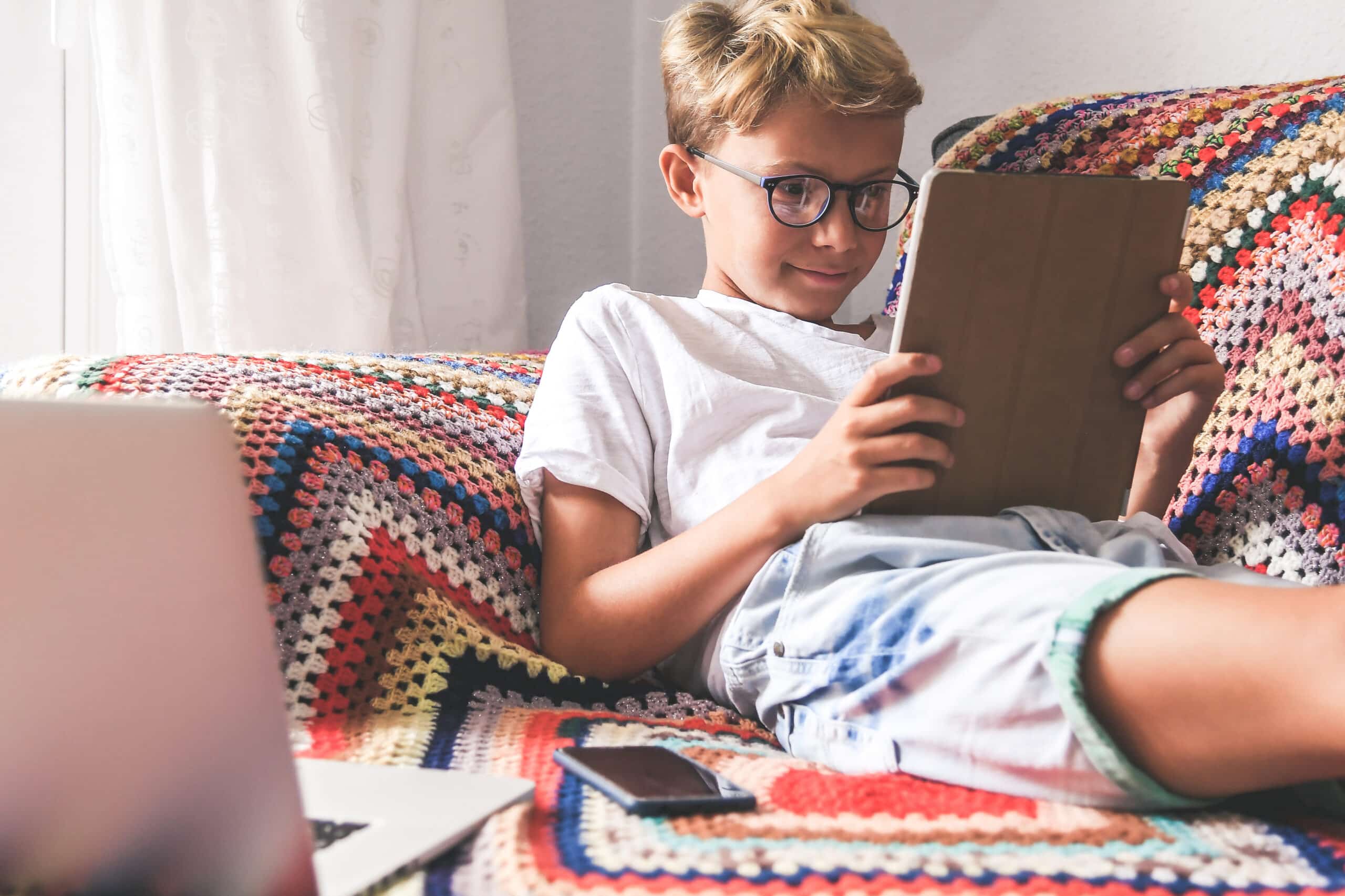 Young boy with glasses sitting on a couch and playing with a tablet